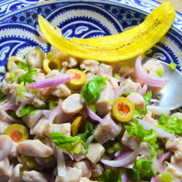 Bowl of chicken salad with sliced olives, red onions, cilantro, and a lemon wedge on a decorative plate.