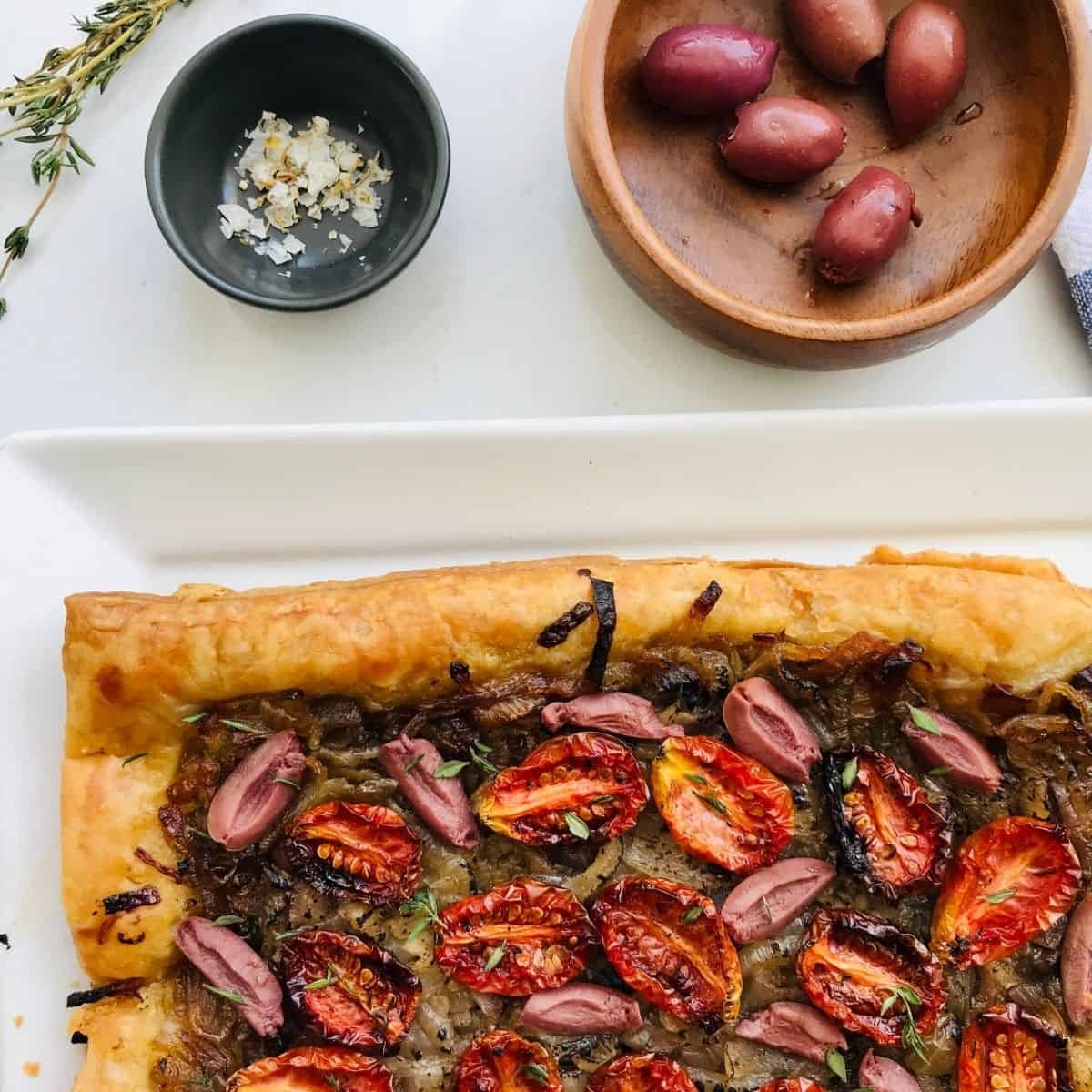 Roasted cherry tomato and caramelized onion tart on a white plate with garlic, fresh herbs, and kalamata olives in wooden bowl, gourmet homemade vegetarian appetizer from Petit Gourmet.