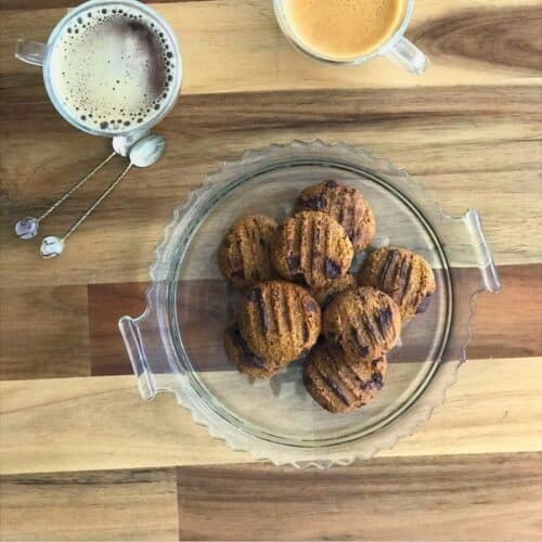 Freshly baked chocolate chip cookies on a glass platter with two cups of coffee on a wooden table, perfect for a gourmet snack or dessert.