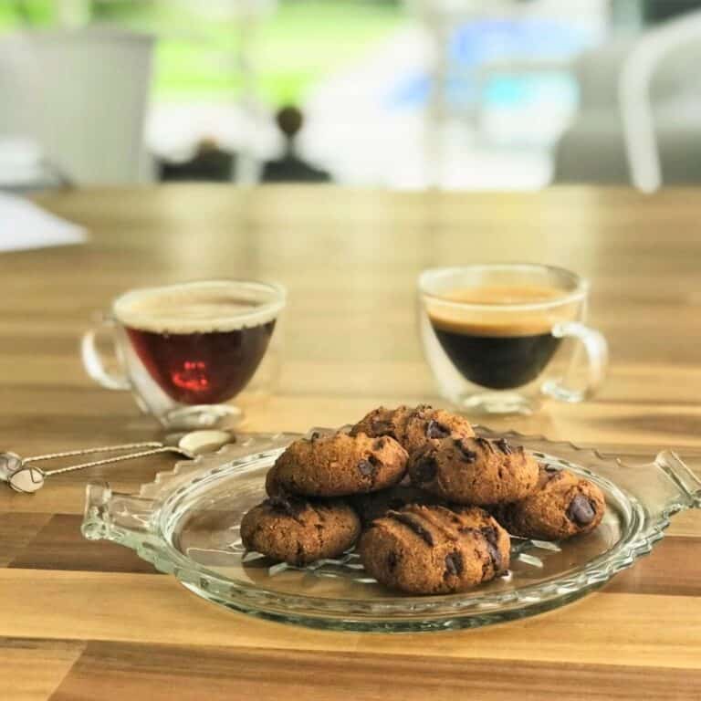 Rich chocolate chip cookies served on a glass platter with two cups of espresso or coffee in the background, perfect for a gourmet dessert or snack.