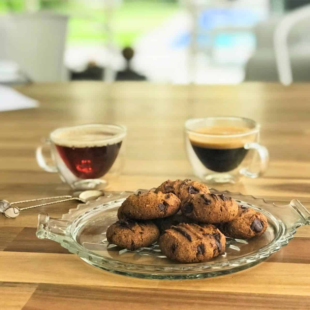 Rich chocolate chip cookies served on a glass platter with two cups of espresso or coffee in the background, perfect for a gourmet dessert or snack.